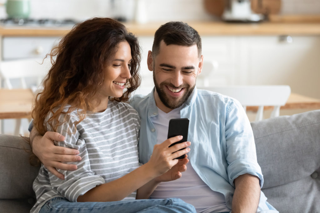 Couple in love resting on comfy couch having fun using smart phone apps, enjoy distant video call, wife showing interesting website to husband, choosing services goods on-line e-commerce usage concept